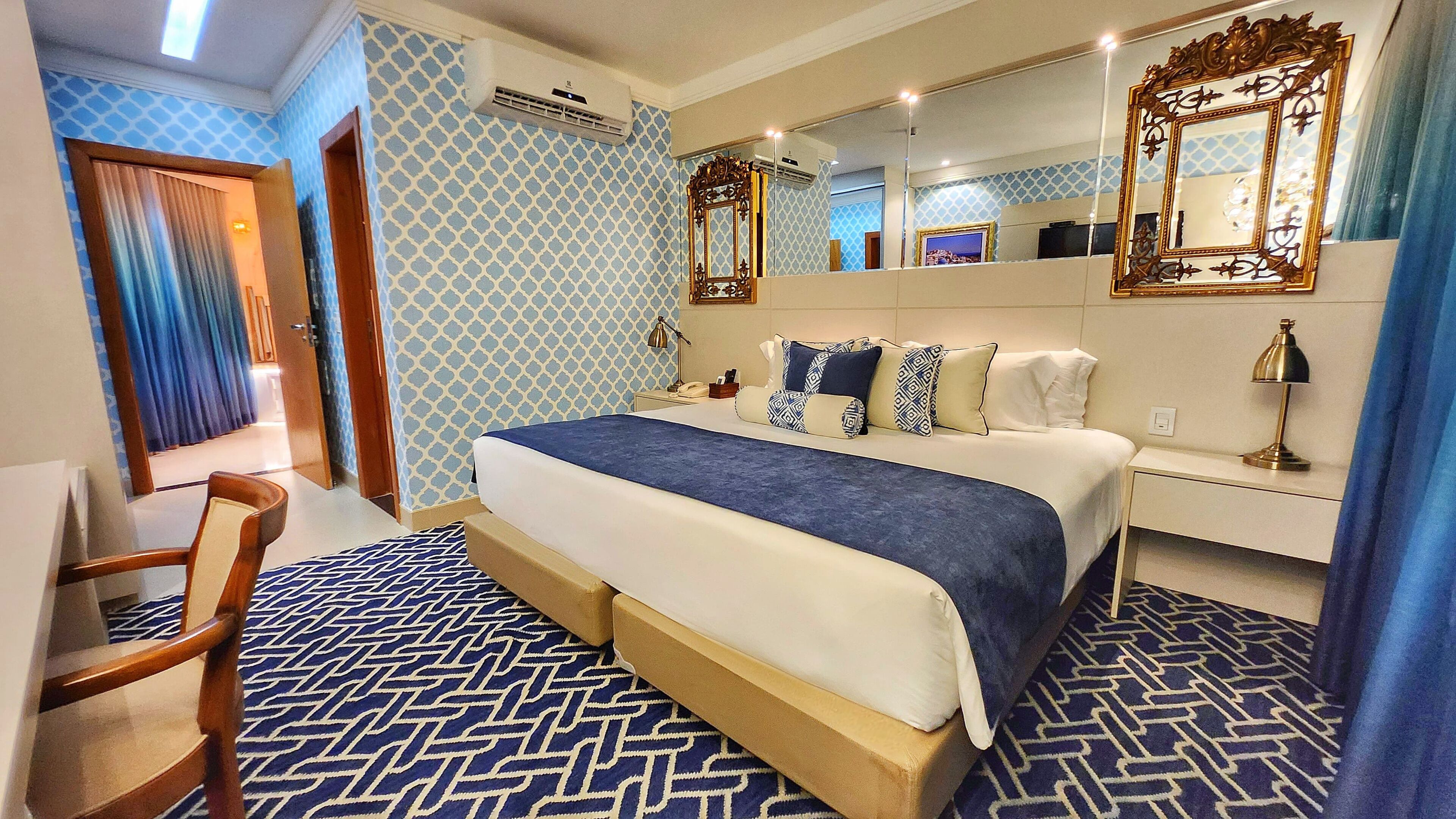 A hotel room with a large bed, a wooden chair, a desk with a lamp, and a large mirror.
