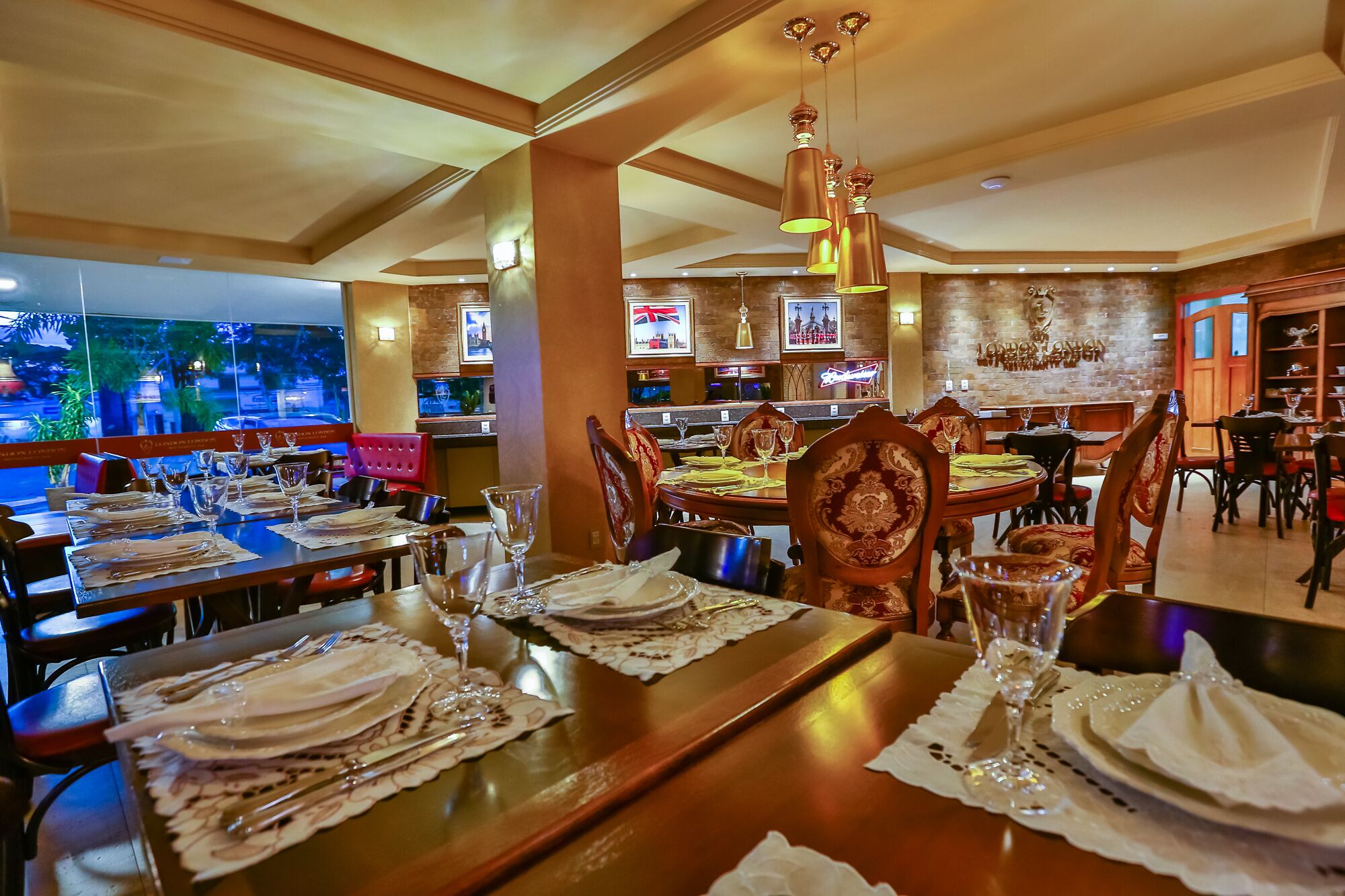 Restaurante com várias mesas de jantar dispostas com toalhas brancas, talheres e copos de vinho. Há luminárias pendentes e uma parede de tijolos com quadros emoldurados.