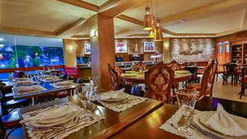 Restaurante com várias mesas de jantar dispostas com toalhas brancas, talheres e copos de vinho. Há luminárias pendentes e uma parede de tijolos com quadros emoldurados.