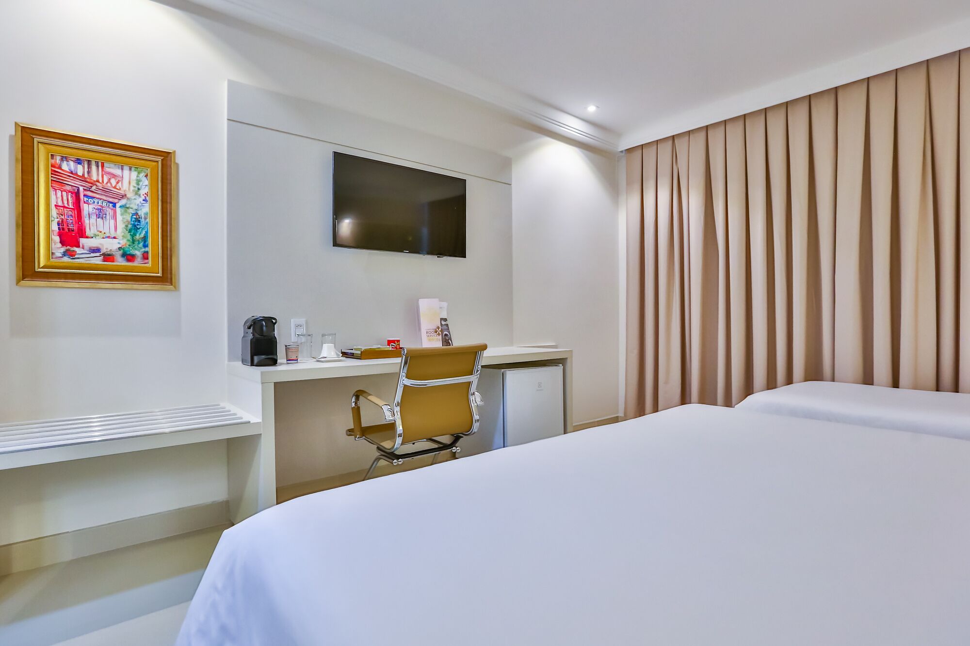 Quarto de hotel com uma cama, uma escrivaninha com cadeira, uma televisão e um quadro na parede.