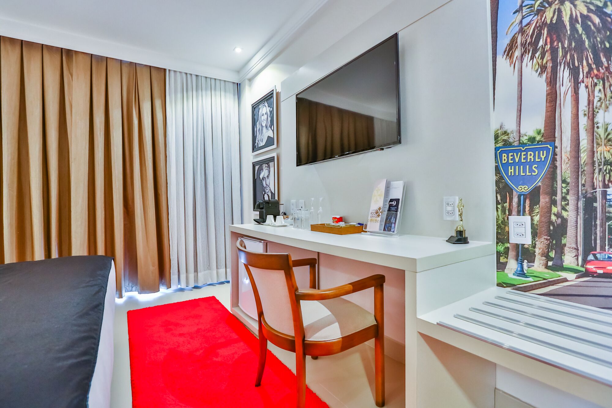 A hotel room with a bed, a desk with a chair, a television, and a poster of Beverly Hills.