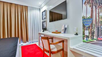 A hotel room with a bed, a desk with a chair, a television, and a poster of Beverly Hills.