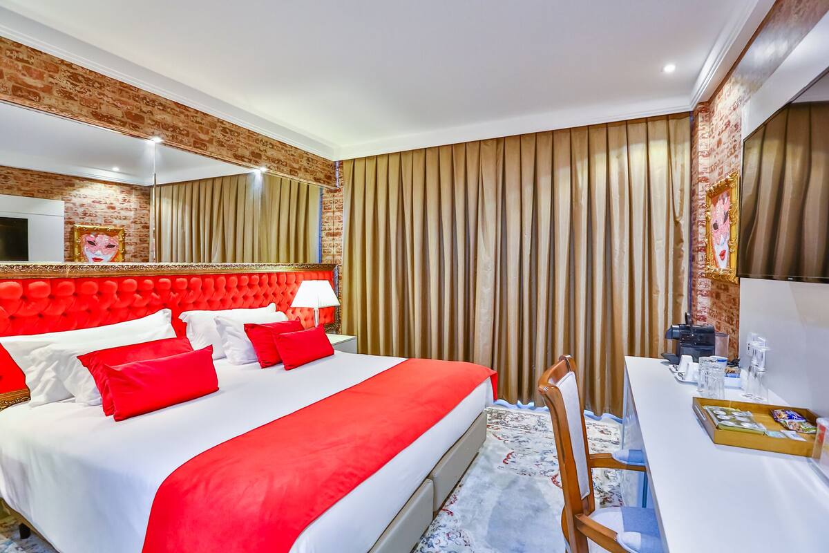 Een hotelkamer met een rode bekleden hoofdeinde, een groot bed met wit en rood beddengoed, een nachtkastje met een lamp en een spiegel boven het bed.