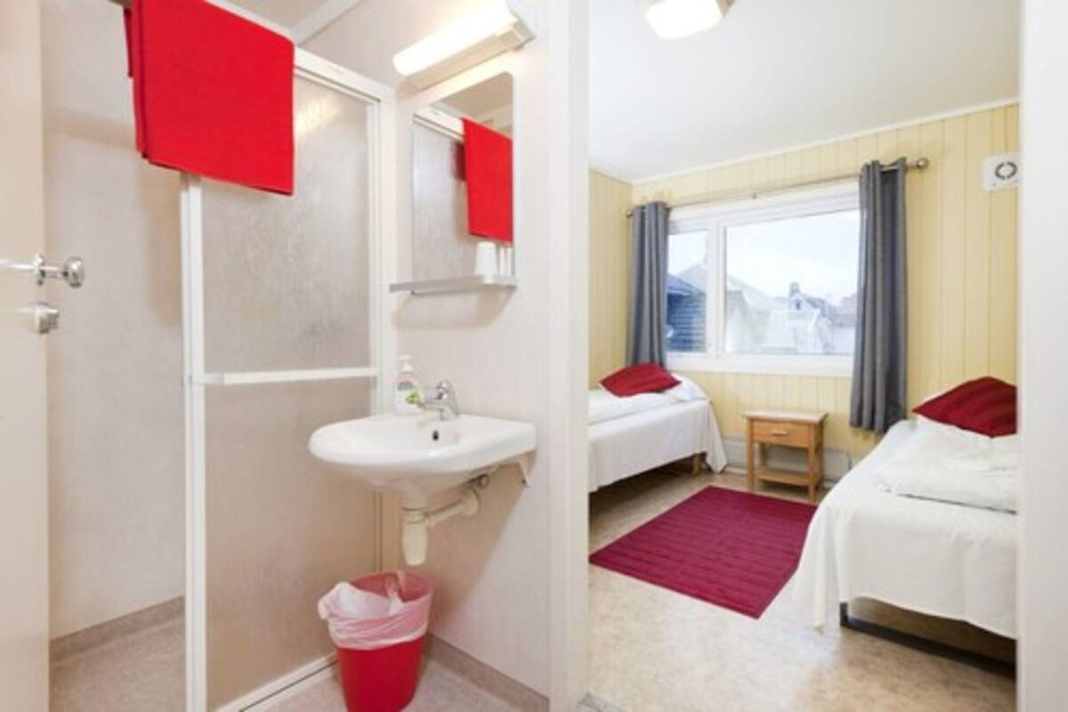Habitación de hotel con cama, mesita de noche, ventana con cortinas, baño con lavabo y espejo, y una toalla roja colgada en la pared.