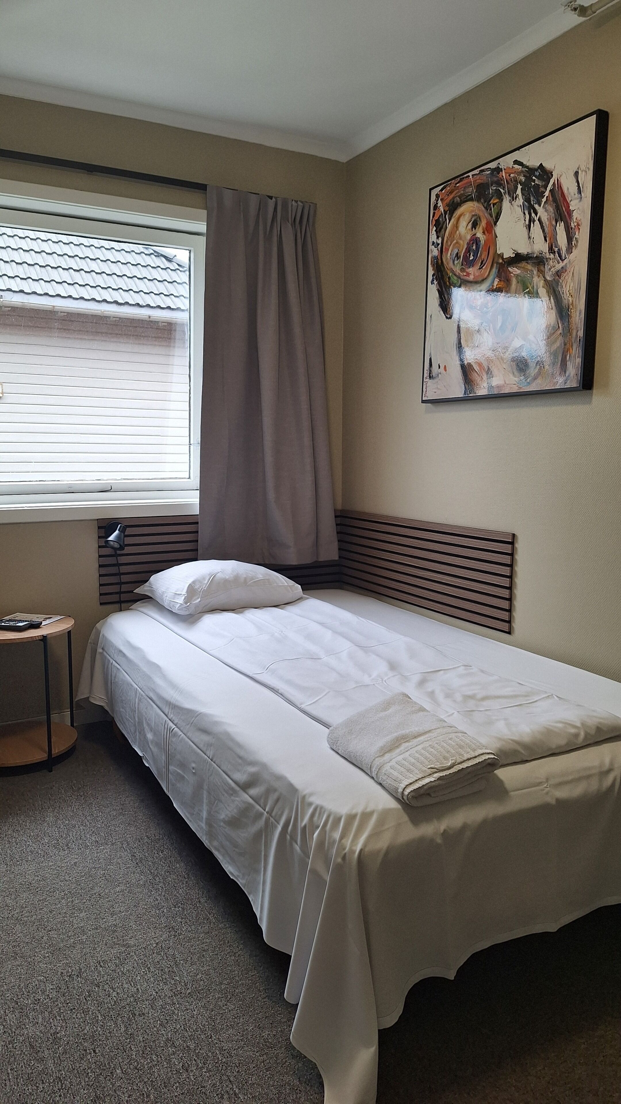 Een net opgemaakt bed in een hotelkamer, met een raam, een bedlampje en een schilderij aan de muur.