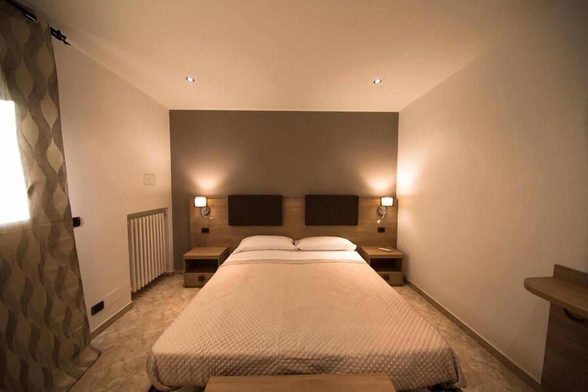 A bedroom with a large bed, two bedside tables with lamps, a radiator, and a window with curtains.