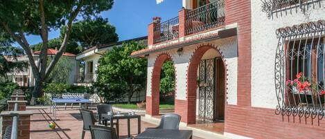 An outdoor patio with tables and chairs, a brick building with arched windows and balconies, and a clear blue sky.