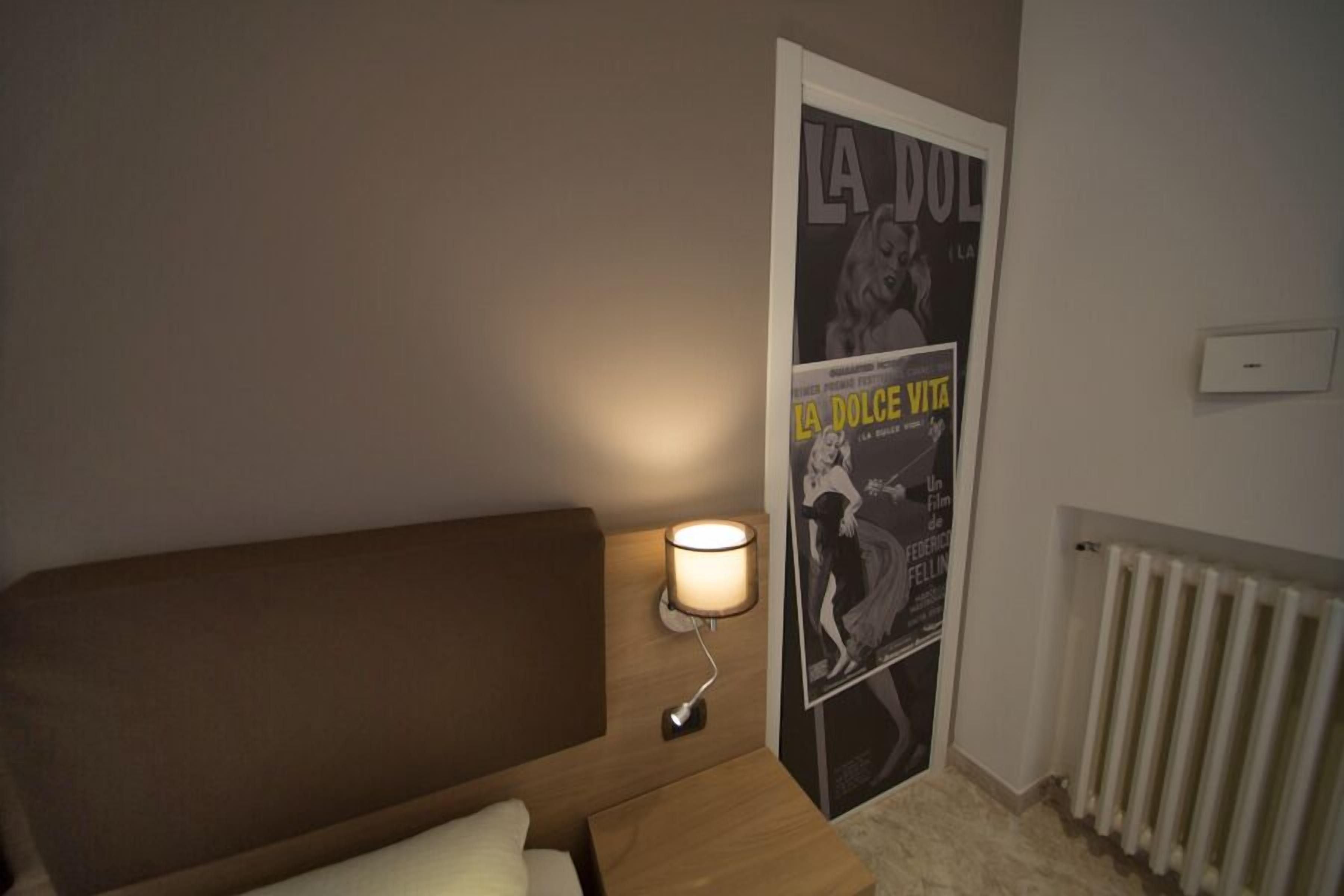 Une chambre d’hôtel avec un lit, une table de chevet avec une lampe, une affiche au mur et un radiateur.