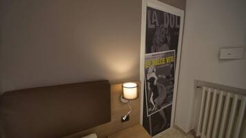 Une chambre d’hôtel avec un lit, une table de chevet avec une lampe, une affiche au mur et un radiateur.