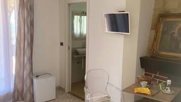 A room with a tiled floor, a television mounted on the wall, a painting above a fireplace, and a door leading to a bathroom.