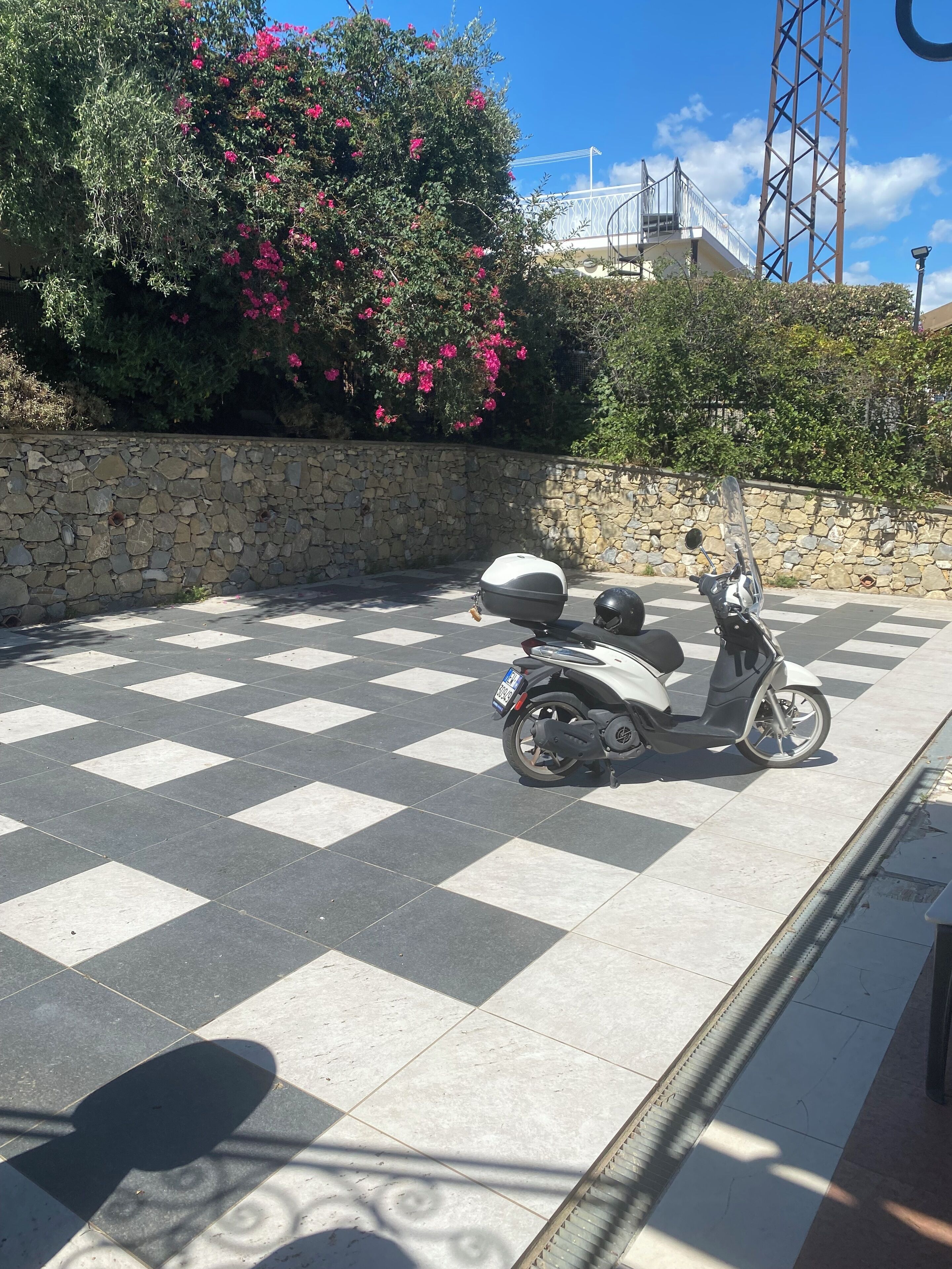 A scooter parked on a checkered pavement with a stone wall and flowering bushes in the background.
