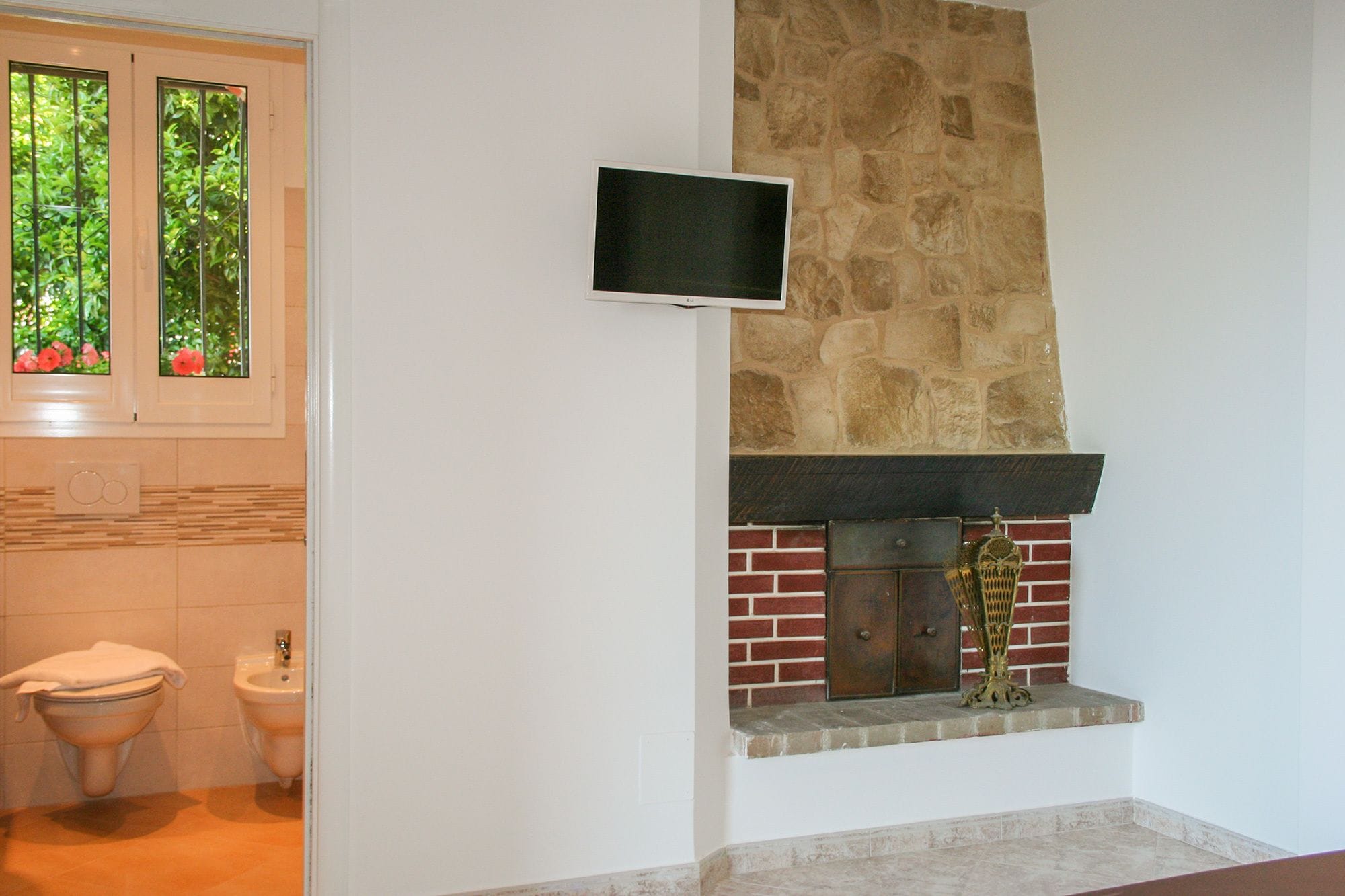 Une salle de bain avec un mur en pierre, un téléviseur fixé au-dessus d’une cheminée et une fenêtre donnant sur un paysage verdoyant.