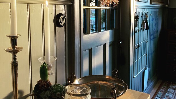 Una habitación con una puerta de madera, una corona colgante, una vela, una bandeja de metal y una planta en maceta.