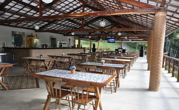 A covered outdoor dining area with wooden tables and chairs, a bar counter, and a television screen. - Quilombo Hotel Fazenda (Uniao dos Palmares)