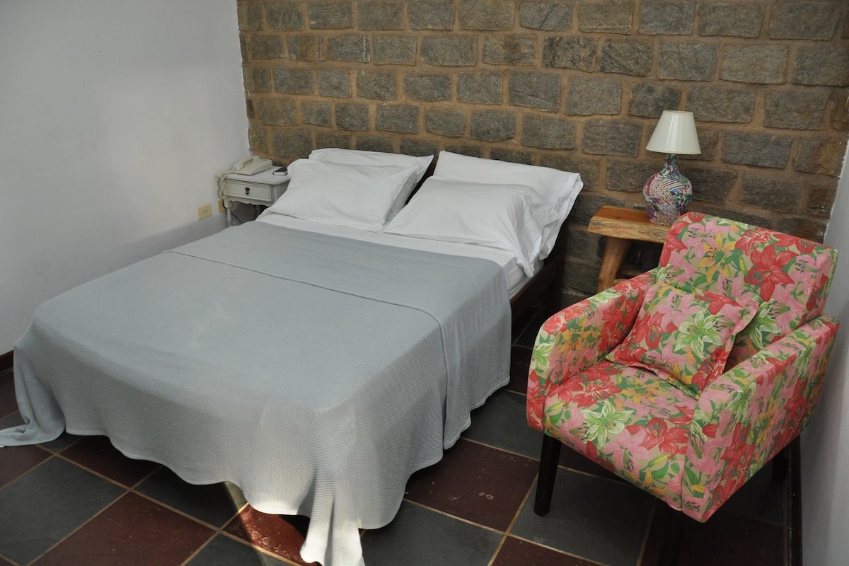A bed with a grey bedspread, a wooden nightstand with a lamp, and a floral armchair.