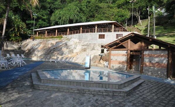 Un área de piscina con sillones de descanso, un pequeño edificio con techo y un muro de piedra.