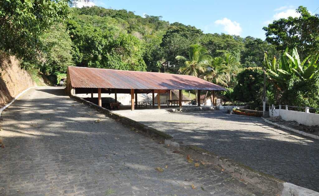 A paved pathway leading to a covered pavilion with a metal roof, surrounded by lush greenery and a hillside.