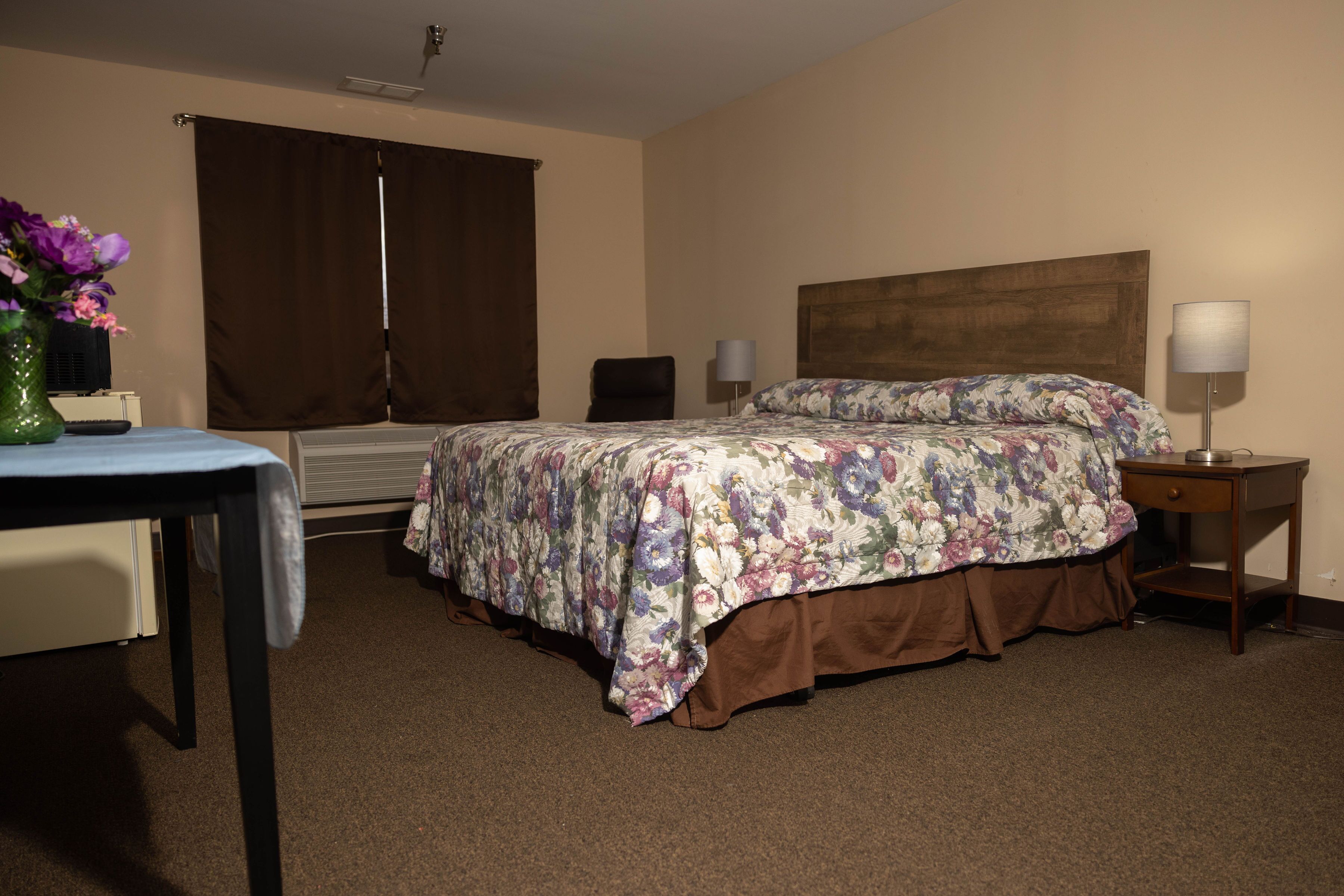 A hotel room with a bed, a desk, a chair, a lamp, and a vase with flowers.