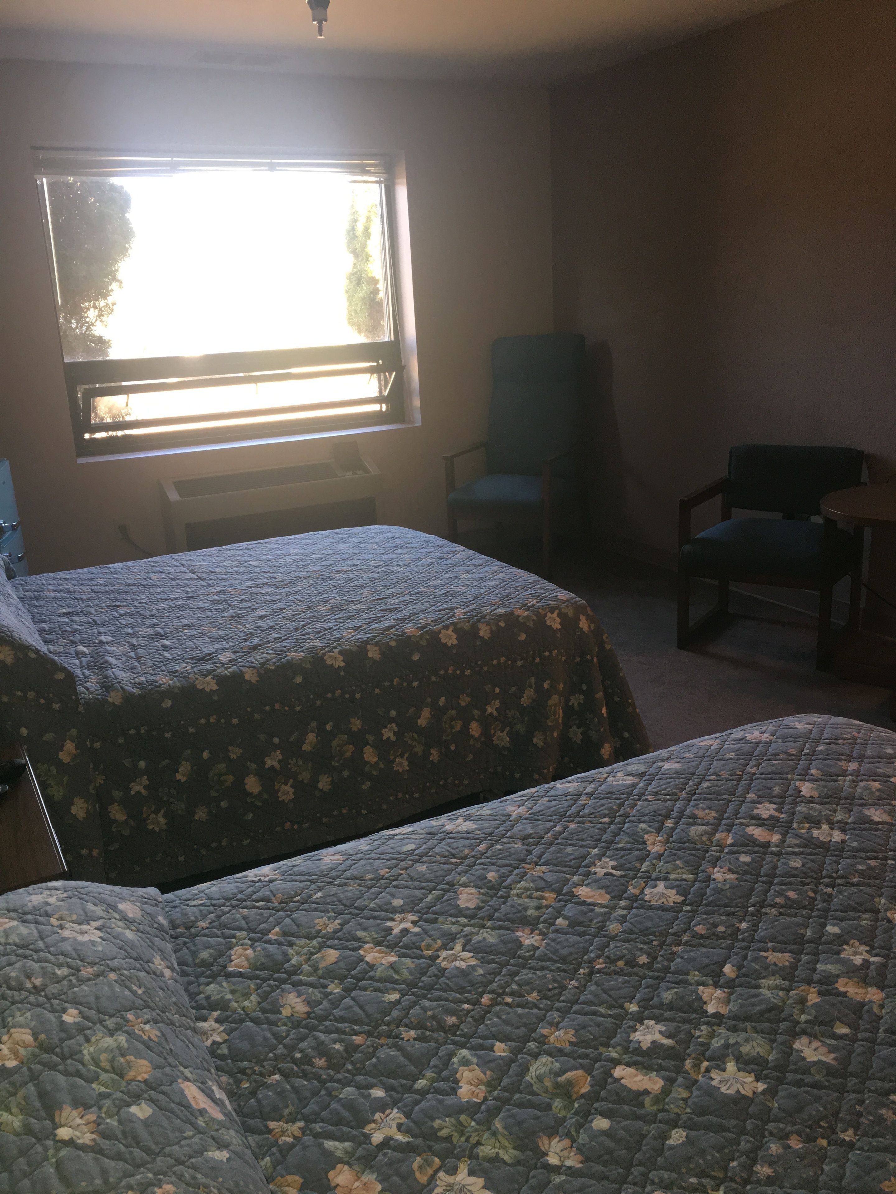 A room with two beds, a chair, a desk, and a window with blinds.