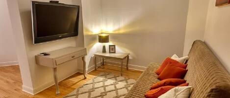 Una sala de estar con un televisor de pantalla plana, una mesita con una lámpara, un sofá con cojines y una alfombra estampada.