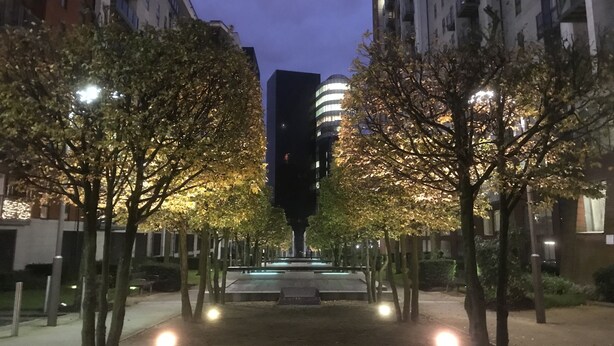 Ein nächtliches Stadtbild mit Bäumen, die einen Weg säumen, einem zentralen Denkmal und modernen Gebäuden im Hintergrund.