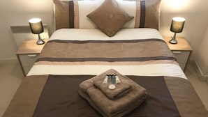 Ein ordentlich bezogenes Bett mit einer braun-beigen Streifenauflage, zwei Nachttischlampen und einem gefalteten Handtuch mit Toilettenartikeln auf dem Bett.