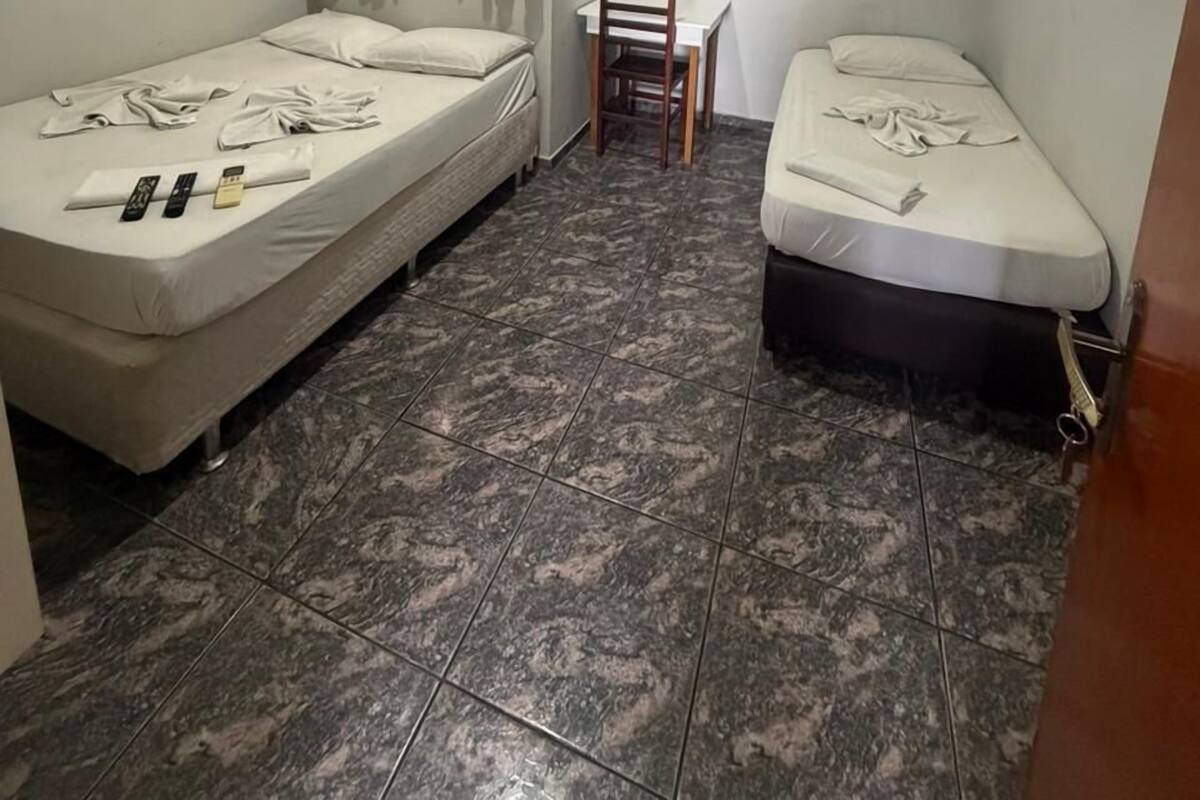 A small room with two single beds, a table, a chair, and an air conditioning unit.