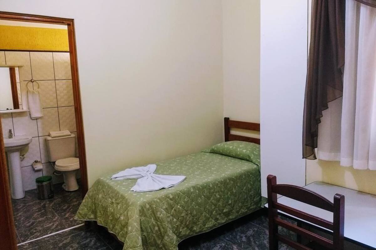 A small, single-bed room with a green bedspread, a wooden headboard, a wooden chair, a small sink, and a toilet.