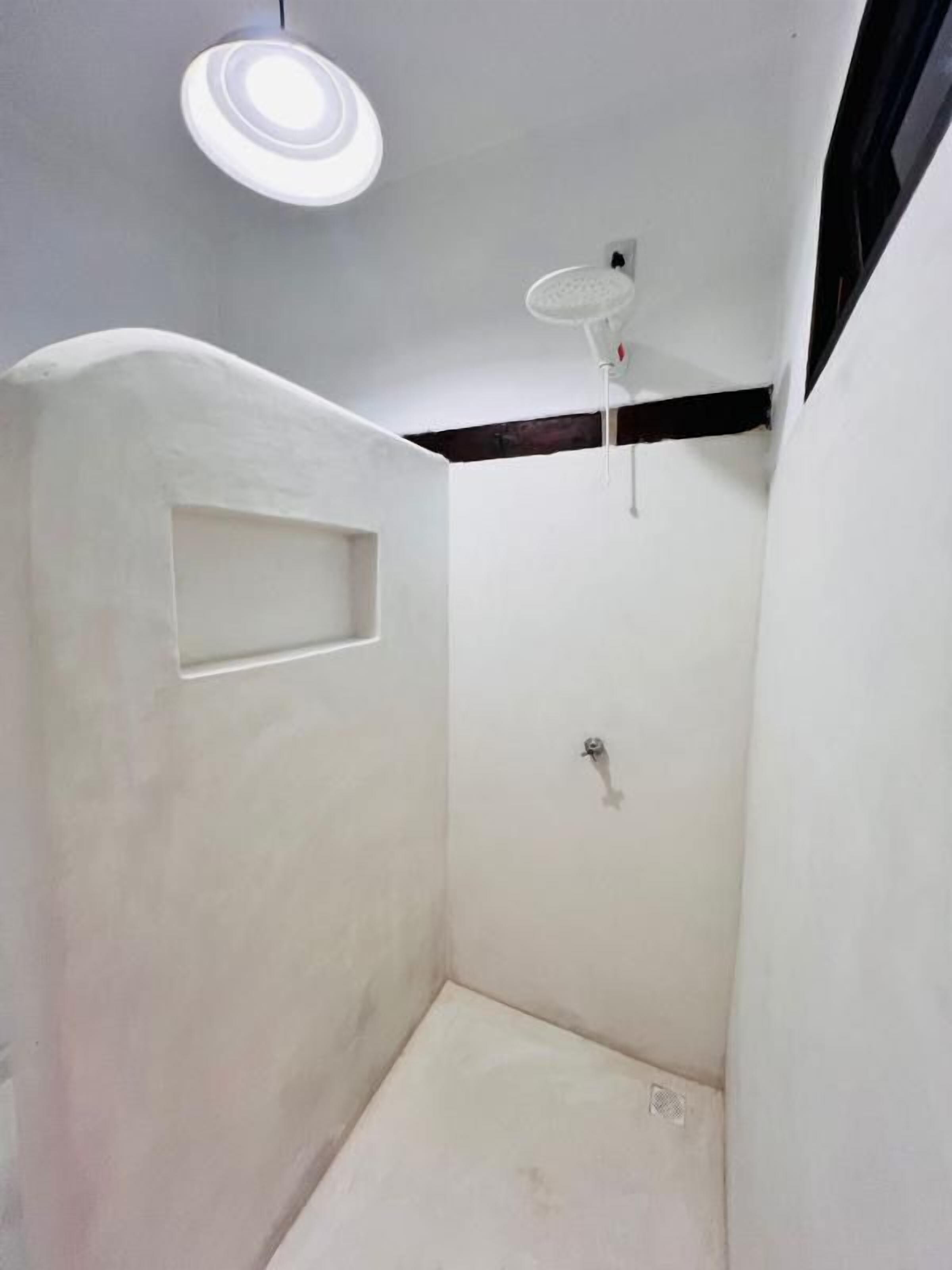 Un pequeño box de ducha blanco, cerrado, con una lámpara en el techo y un grifo empotrado en la pared.