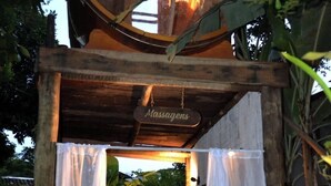 A massage parlor with a wooden archway, a sign reading 'Massagens,' and a person attending to a client on a bed. - Pousada Raízes do Brasil (Porto Seguro)