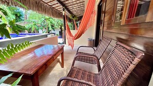 A wooden bench and wicker chairs under a thatched roof, with a pool and greenery in the background.