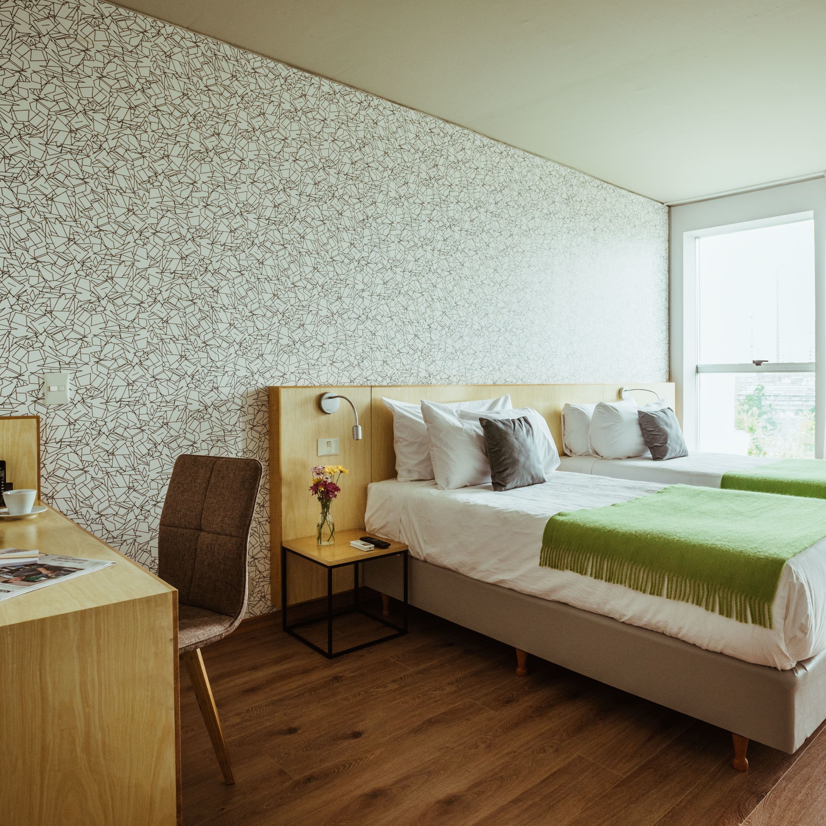 A hotel room with a wooden desk, a chair, a bedside table with a vase of flowers, a bed with pillows and a green throw blanket, and a patterned wallpaper.