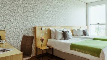 A hotel room with a wooden desk, a chair, a bedside table with a vase of flowers, a bed with pillows and a green throw blanket, and a patterned wallpaper.