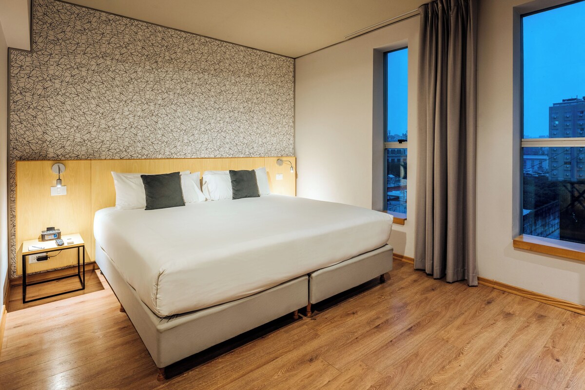 A modern hotel room with a large bed, wooden flooring, a city view through the window, and a patterned wall.