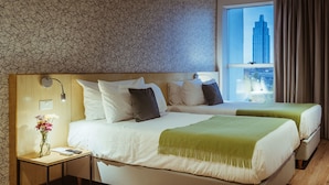 Habitación de hotel con dos camas, una mesita de noche, una lámpara y vista a un paisaje urbano a través de un ventanal grande.