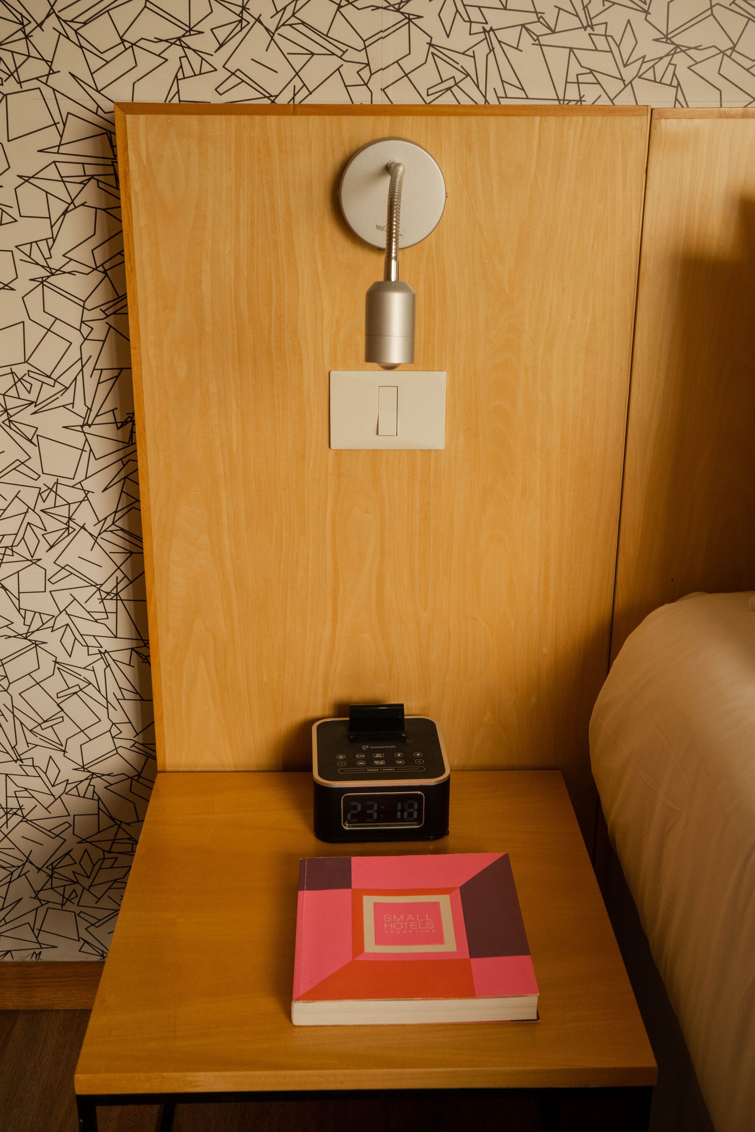 A wooden bedside table with a book titled "Small Hotels" and a digital clock.