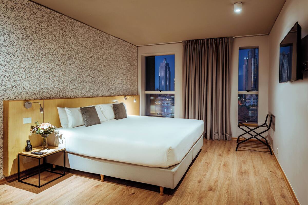 A modern hotel room with a large bed, a bedside table with a lamp, a small table with a vase of flowers, and a chair. The room has a city view through two windows.