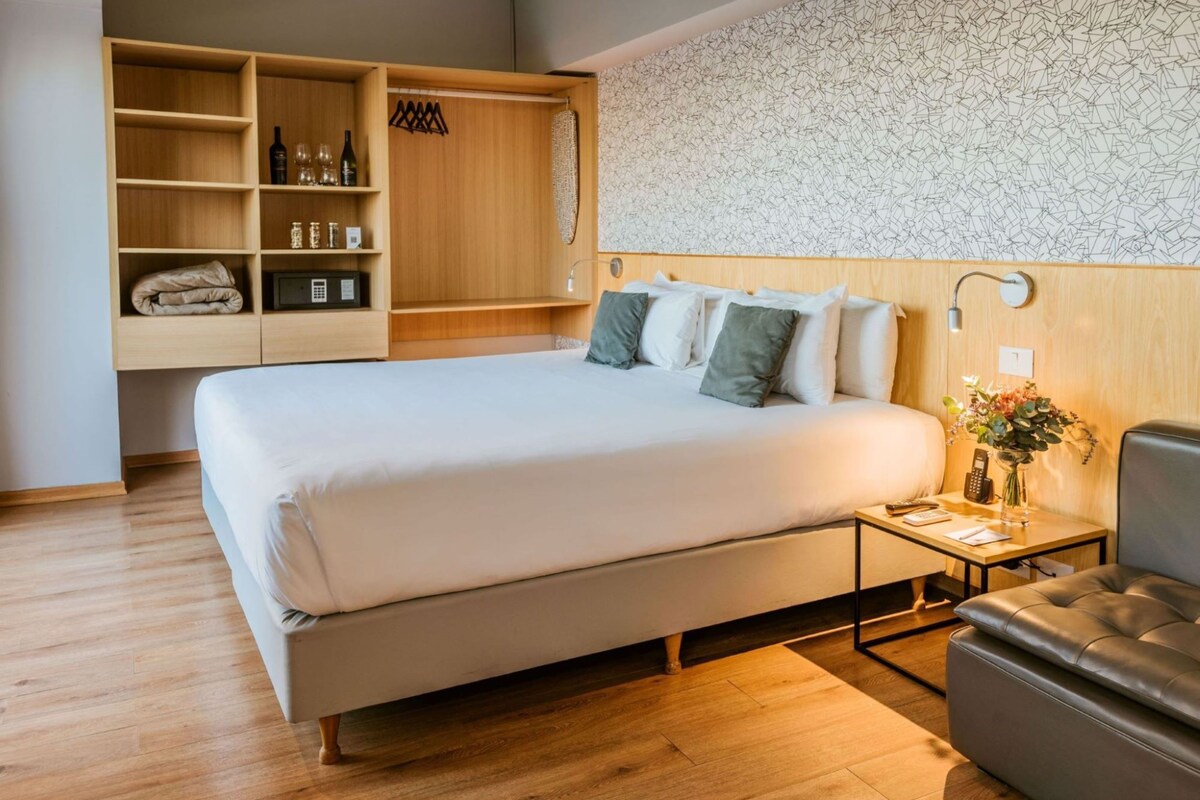 A modern hotel room with a large bed, wooden wardrobe, bedside table, and a small seating area.