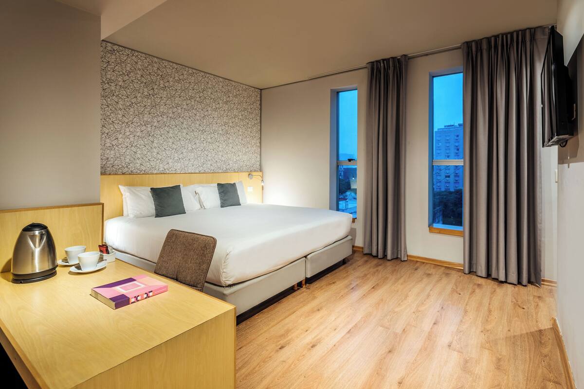 A hotel room with a bed, a desk with a kettle, a cup, and a book. There is a window with curtains and a view of buildings outside.