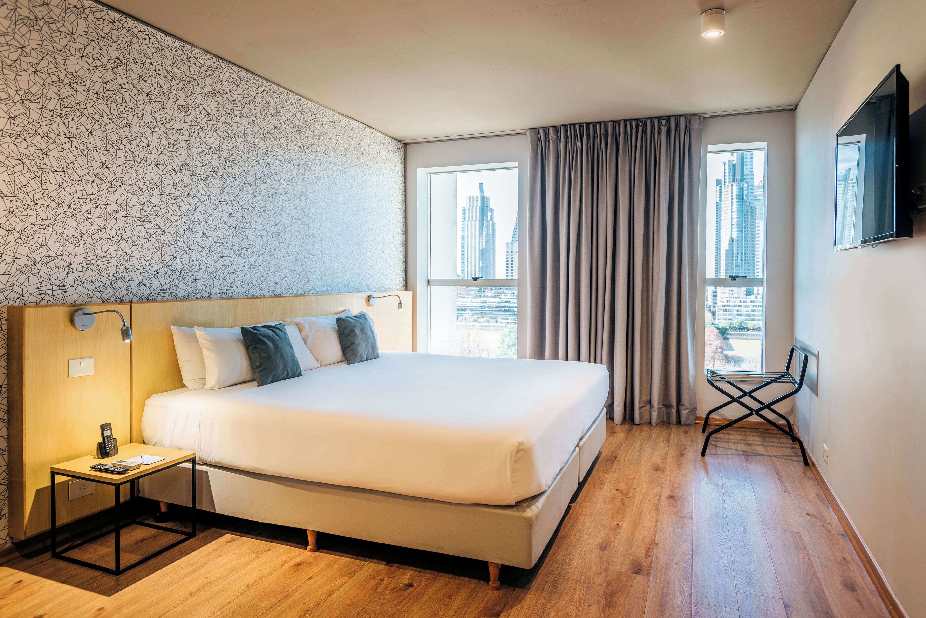 Quarto de hotel com uma cama grande, uma mesa com telefone e vista para a paisagem urbana pela janela.