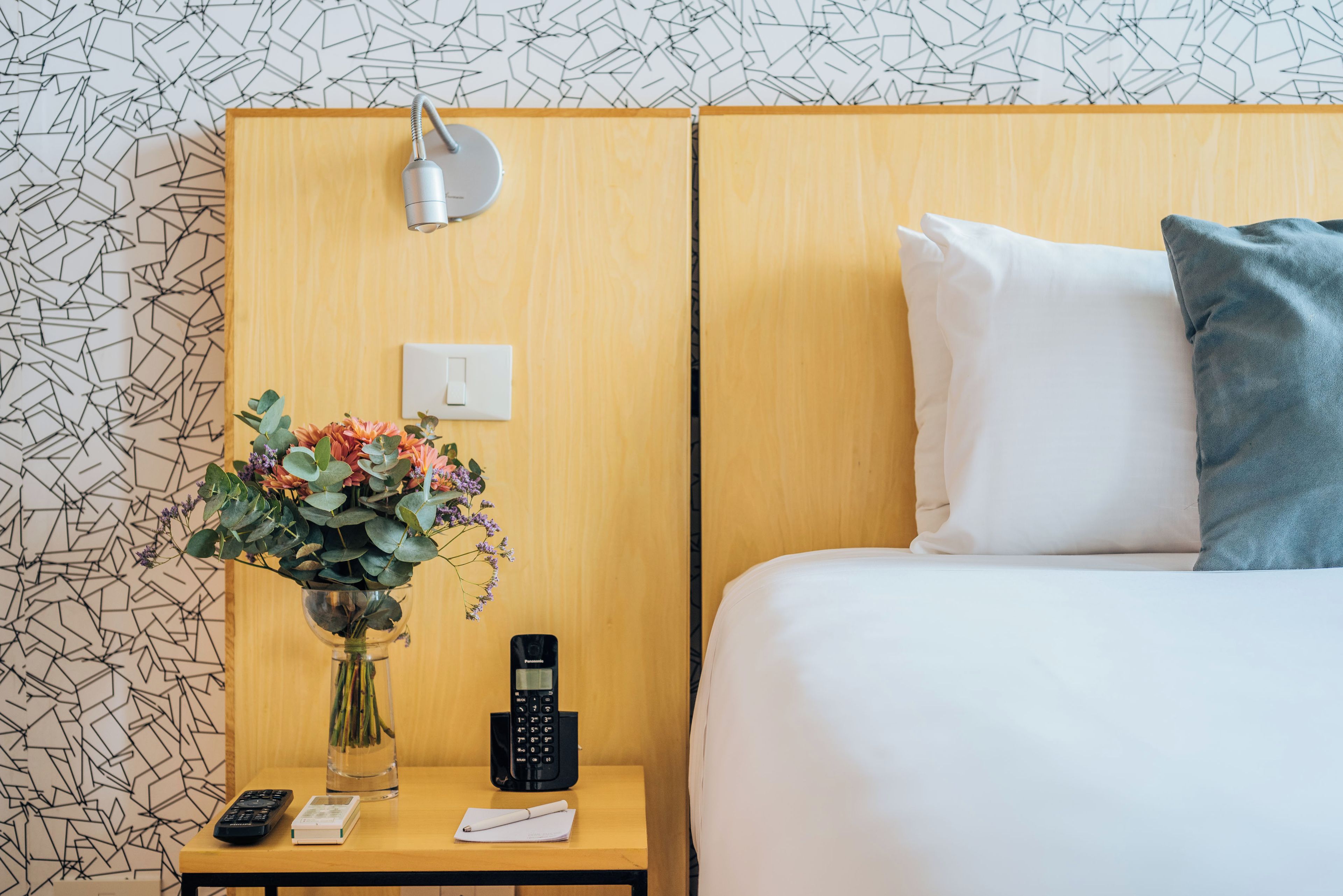 A hotel room with a wooden headboard, a bedside table with a vase of flowers, a remote control, a phone, and a pillow on the bed.