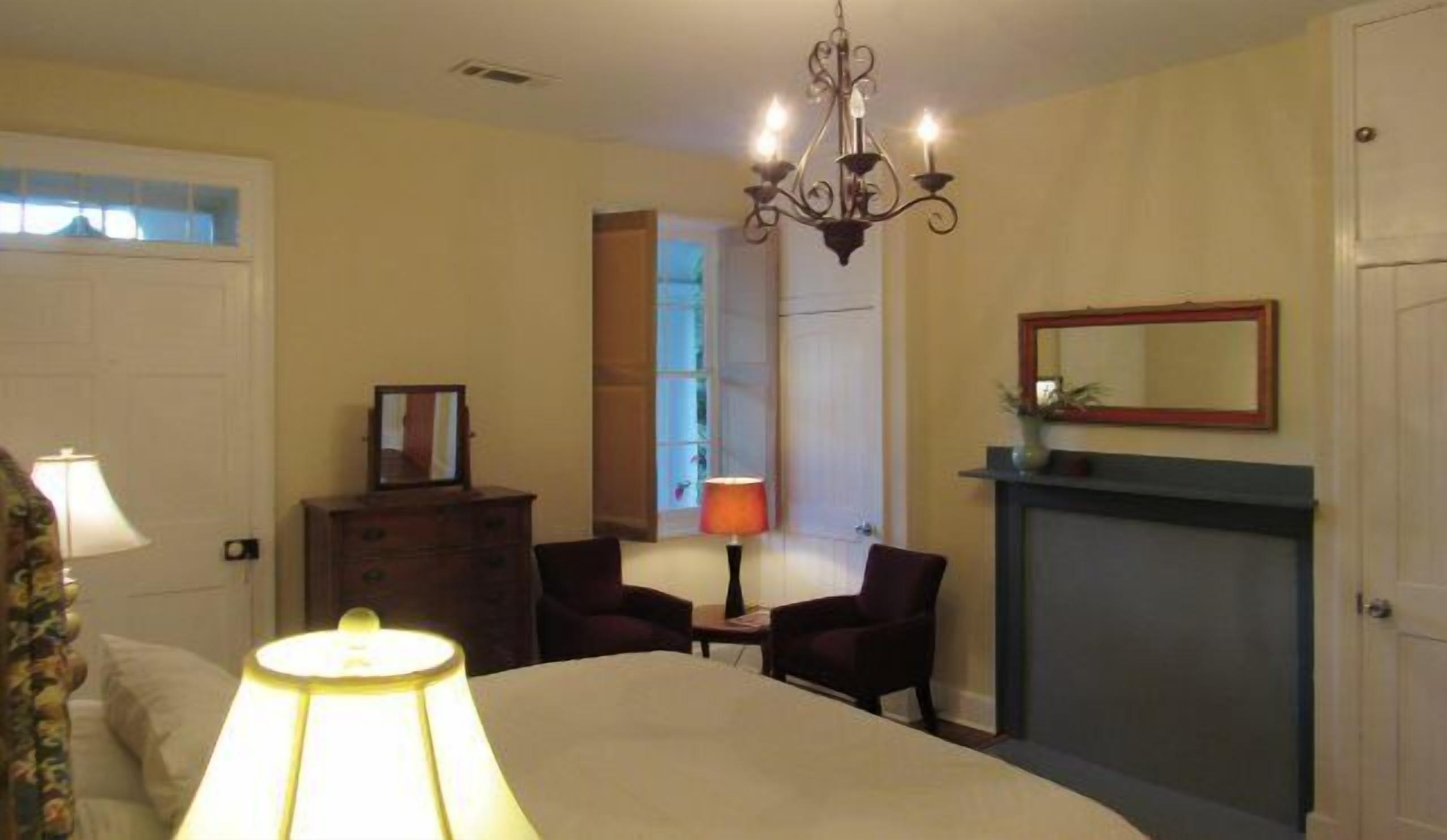 A bedroom with a bed, a dresser, a chair, a lamp, a mirror, and a fireplace.