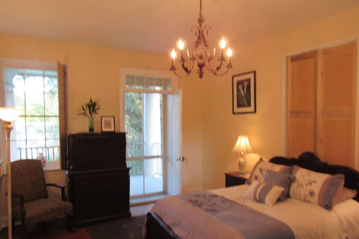 Une chambre à coucher comprenant un lit, une commode, une chaise, une lampe, un lustre et une porte donnant sur un balcon.