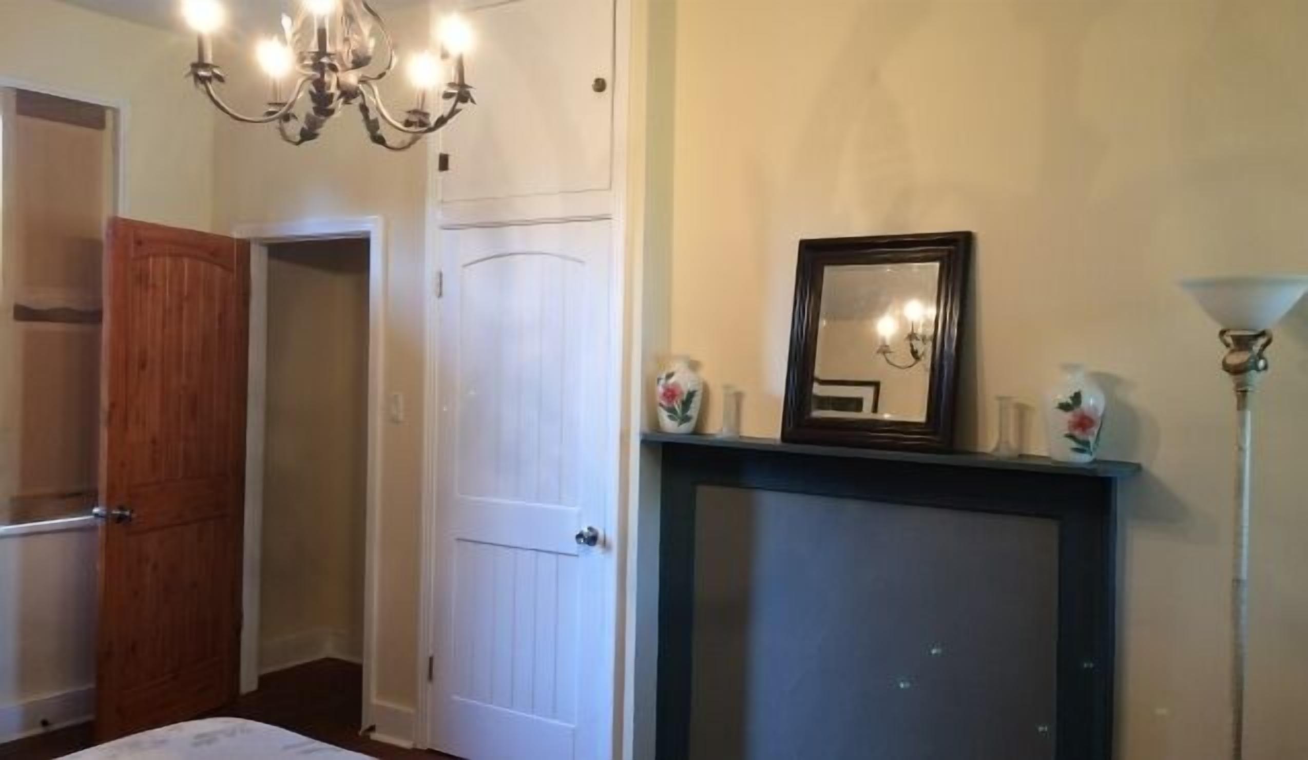 Un dormitorio con cama, un armario de madera, una puerta blanca, una araña, un espejo y una chimenea.