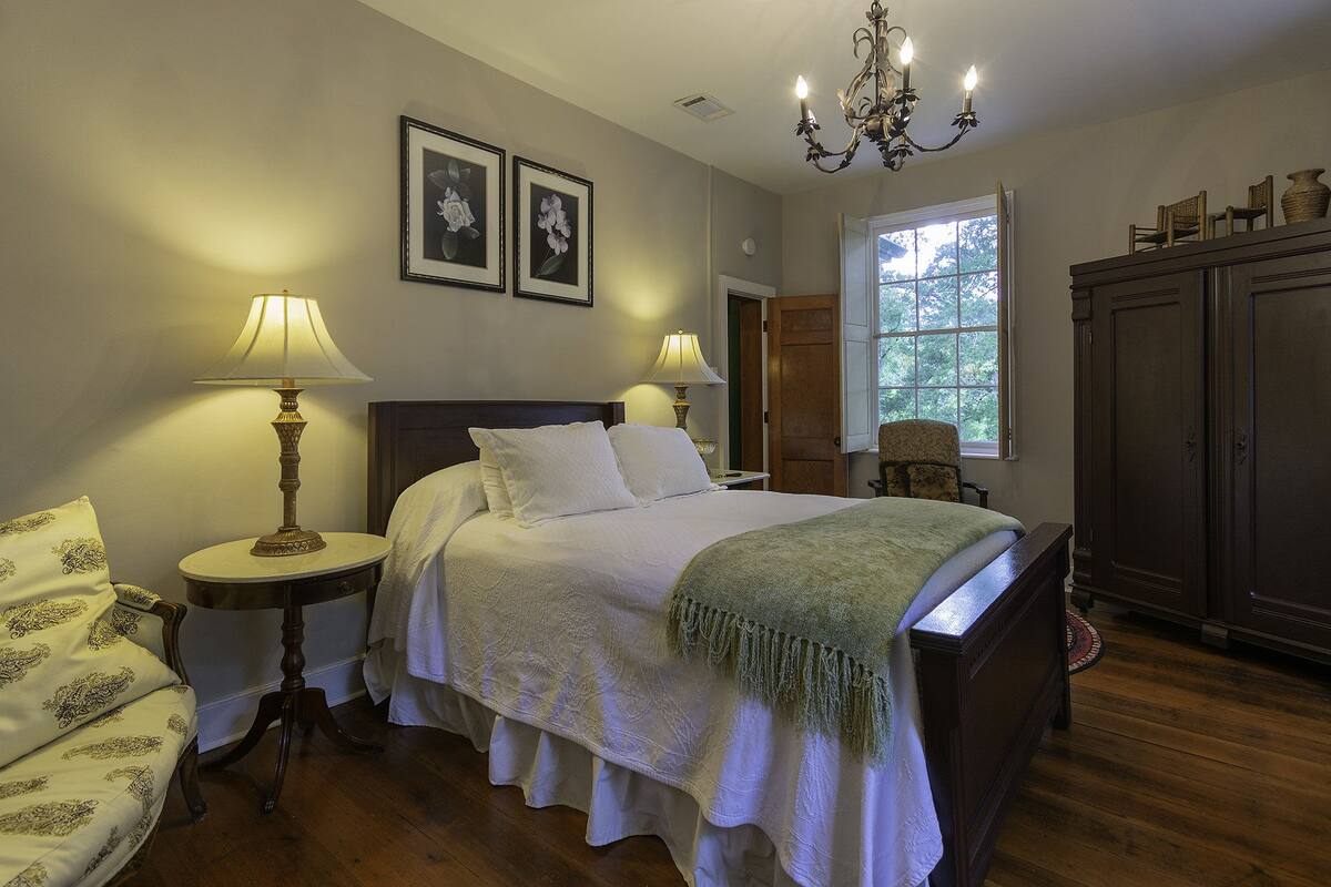 Une chambre à coucher comprenant un lit, deux lampes de chevet, une chaise, une armoire et une fenêtre donnant sur des arbres.
