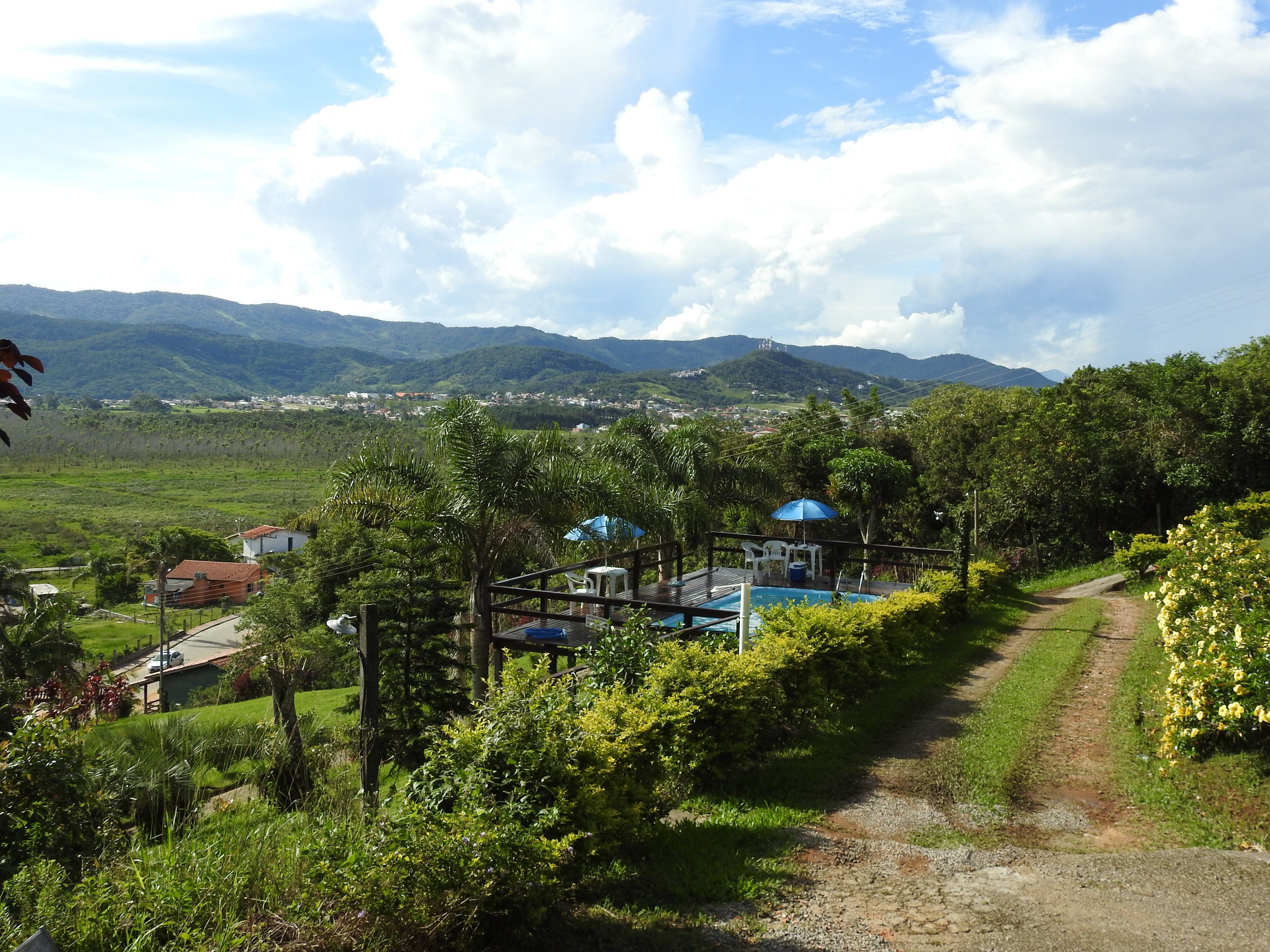 A rural landscape with a dirt path, a small pool area with a blue umbrella, and a view of distant mountains and a village.