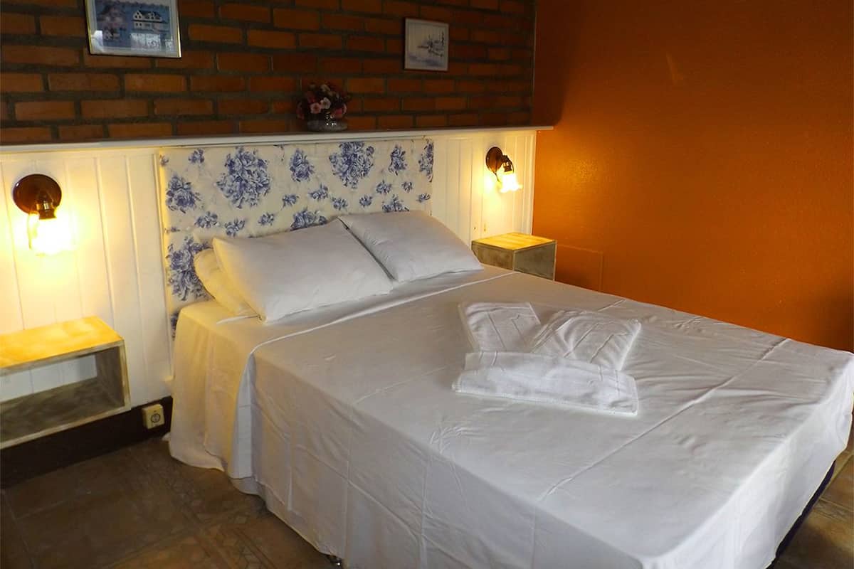 A neatly made bed with white linens and a folded towel, two bedside tables with lamps, a floral-patterned headboard, and a brick wall with framed pictures.