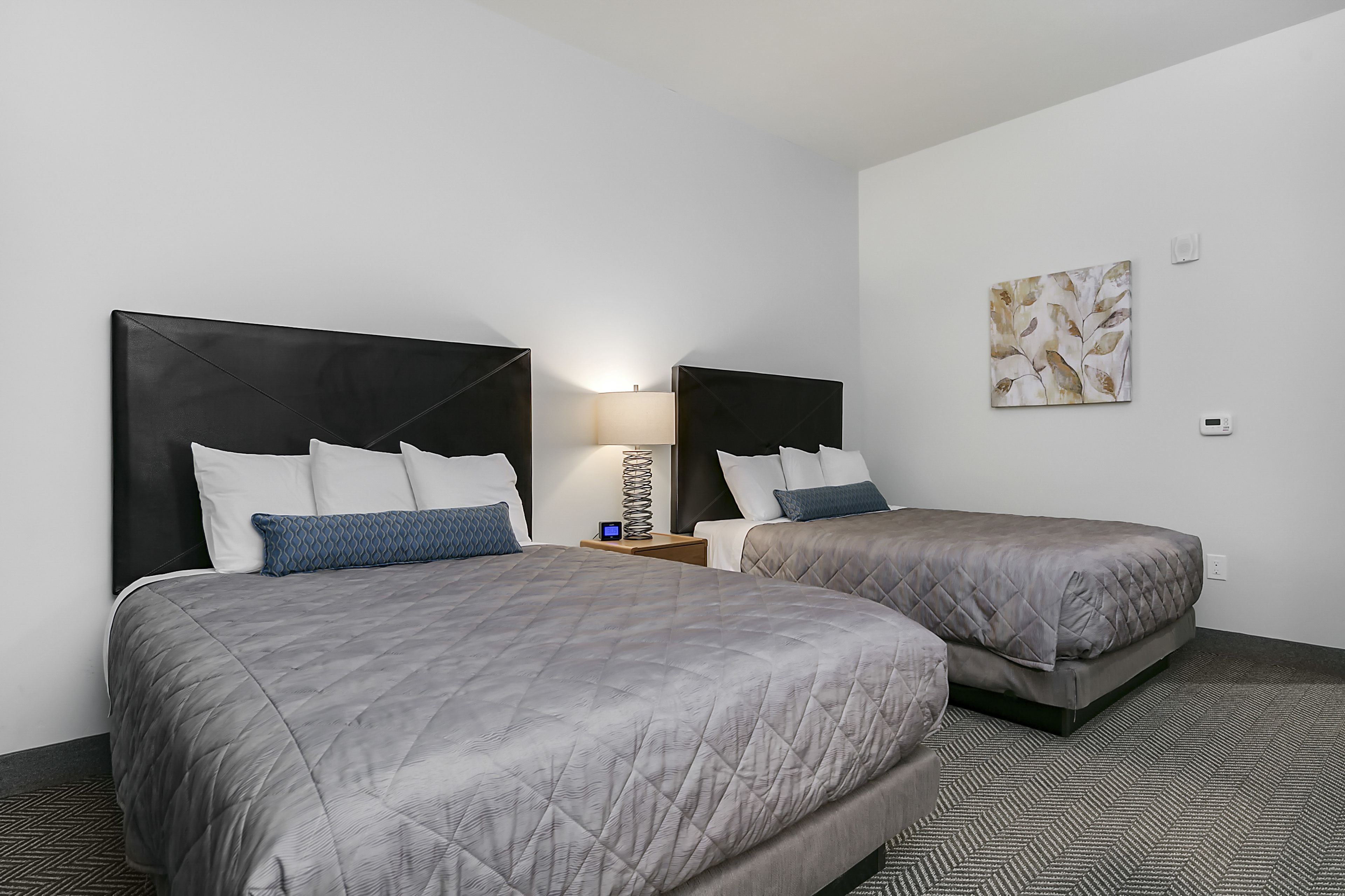 A hotel room with two beds, a nightstand, a lamp, and a wall-mounted thermostat.