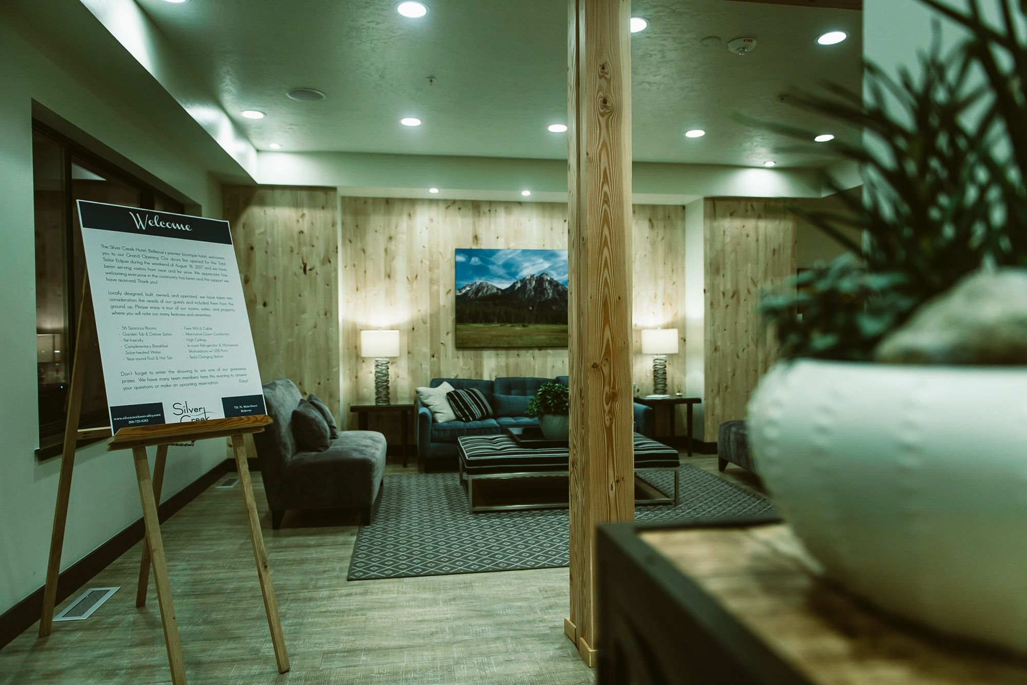 A hotel lobby with a reception desk, a large potted plant, a sofa, a coffee table, and a painting of mountains on the wall.