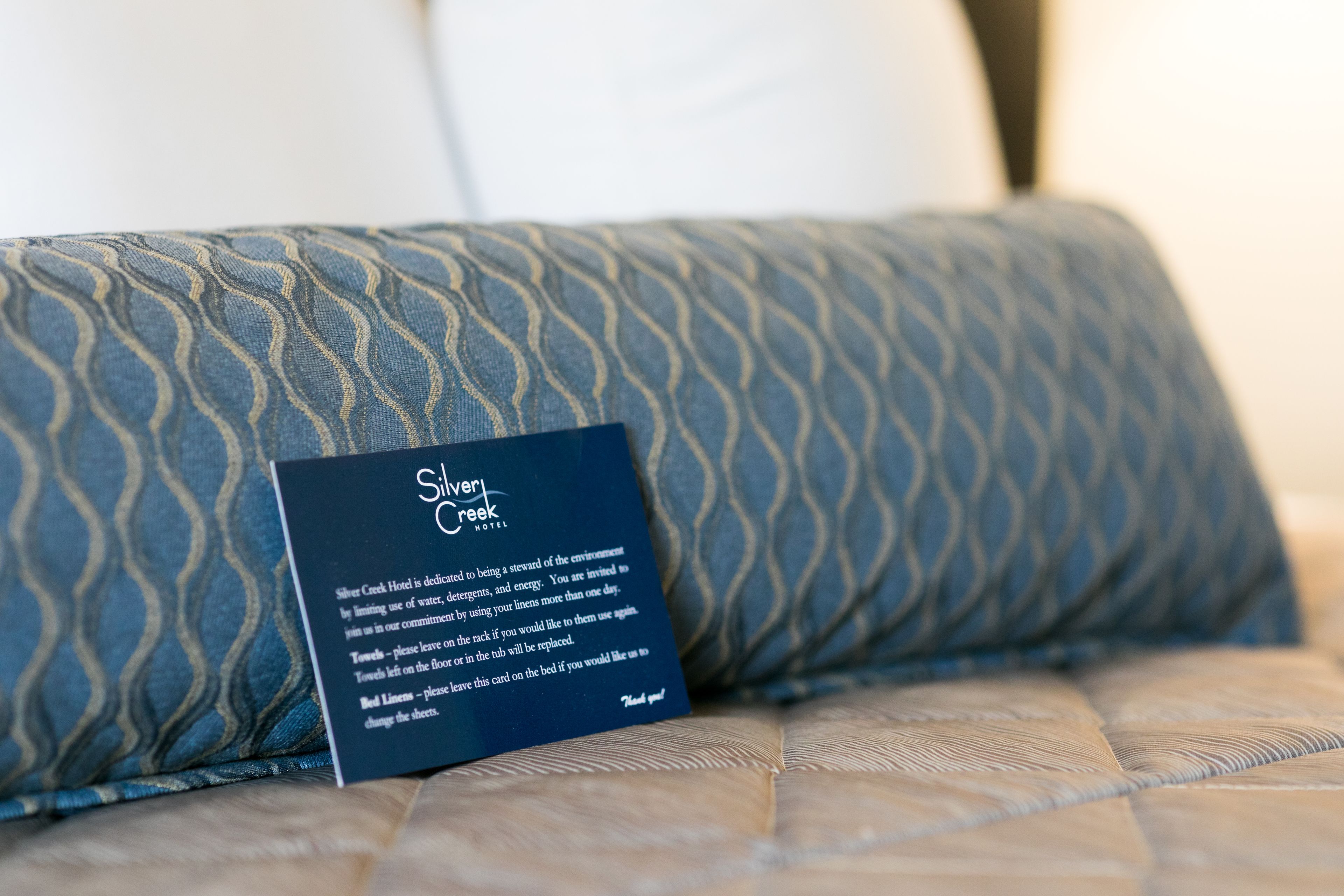 A hotel room with a bed, a pillow with a blue and gold pattern, and a card on the pillow with text.